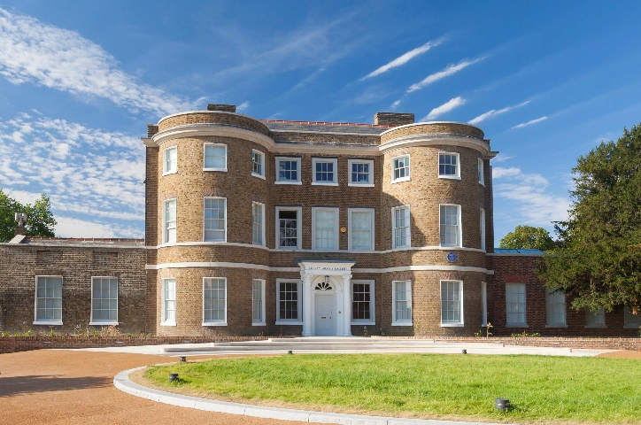 Exterior image of William Morris Gallery. It is a sunny day and light is shining on the left side of the Gallery. There are blue skies with clouds visible. The grass circle in front of the Gallery can also be seen.