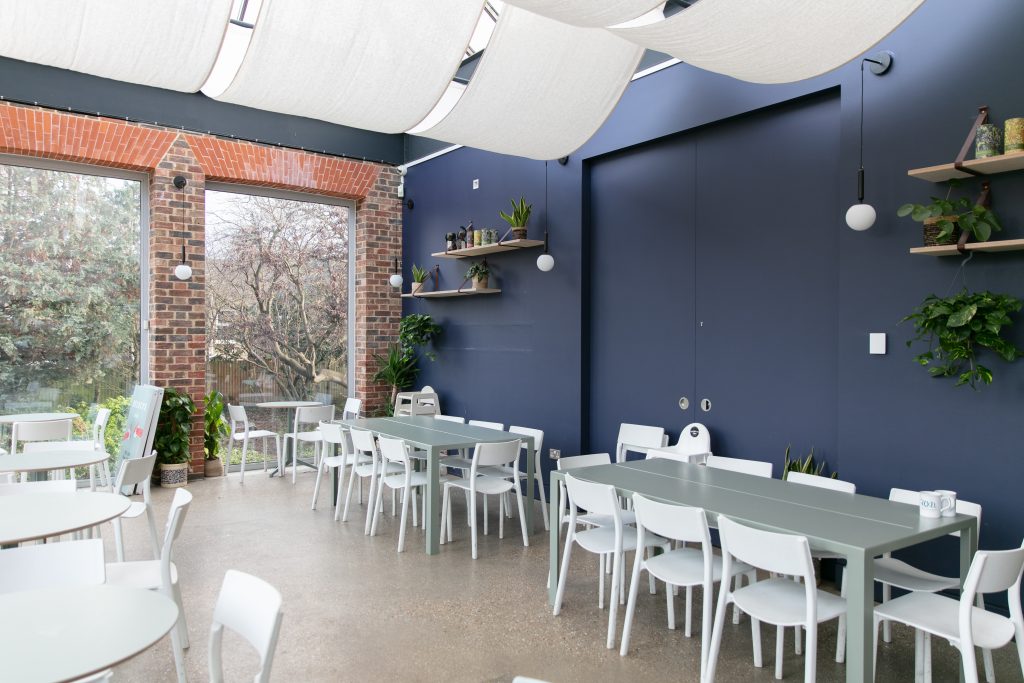 Image of the cafe at William Morris Gallery. White tables and chairs can be seen, with dark blue walls and large windows showing Lloyd Park. Fabric drapes the ceiling of this space.