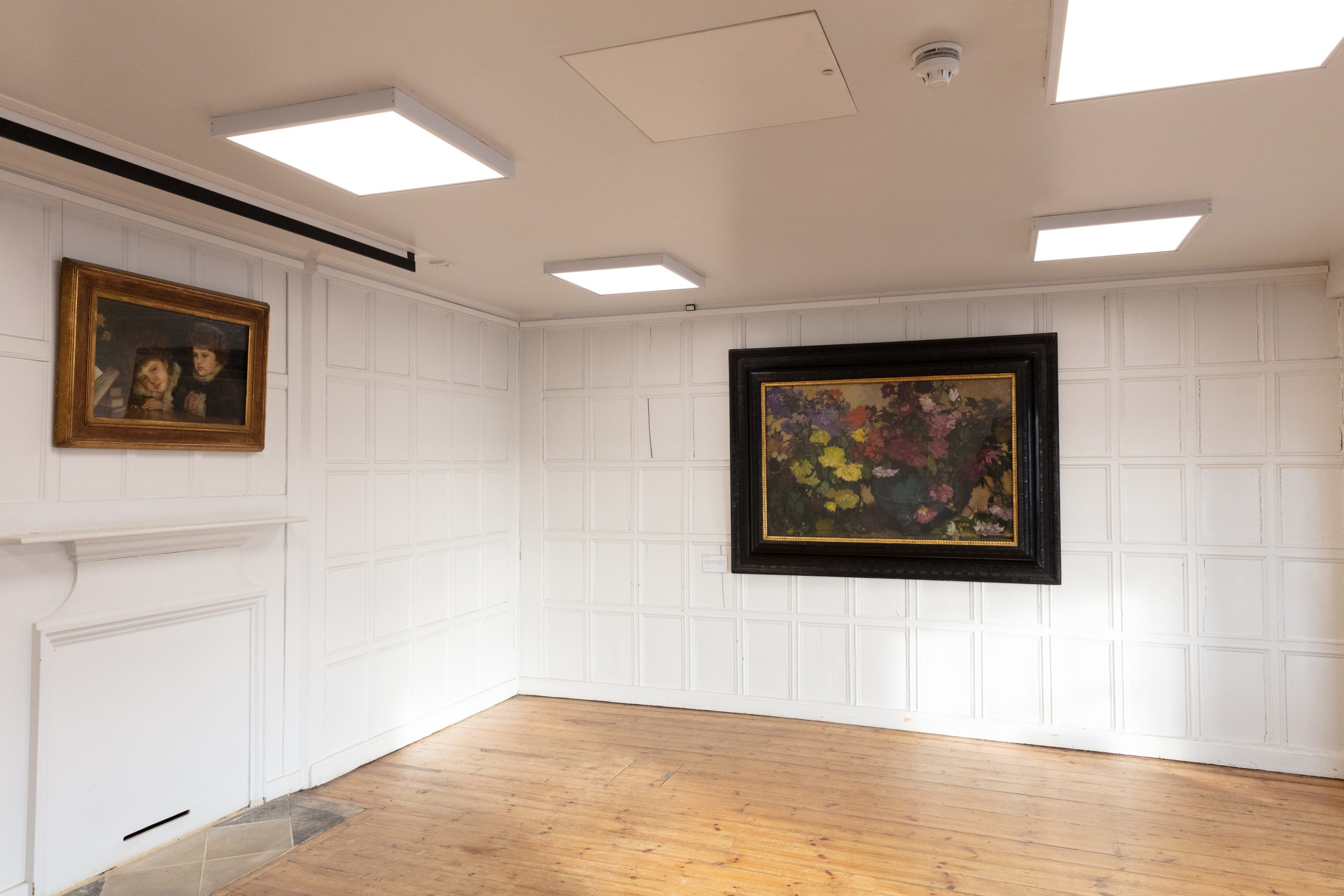 The Acanthus room at William Morris Gallery. The floors are wooden and we have three windows to the right, with sun shining through them. The walls of the room are painted white. There are paintings on the walls.