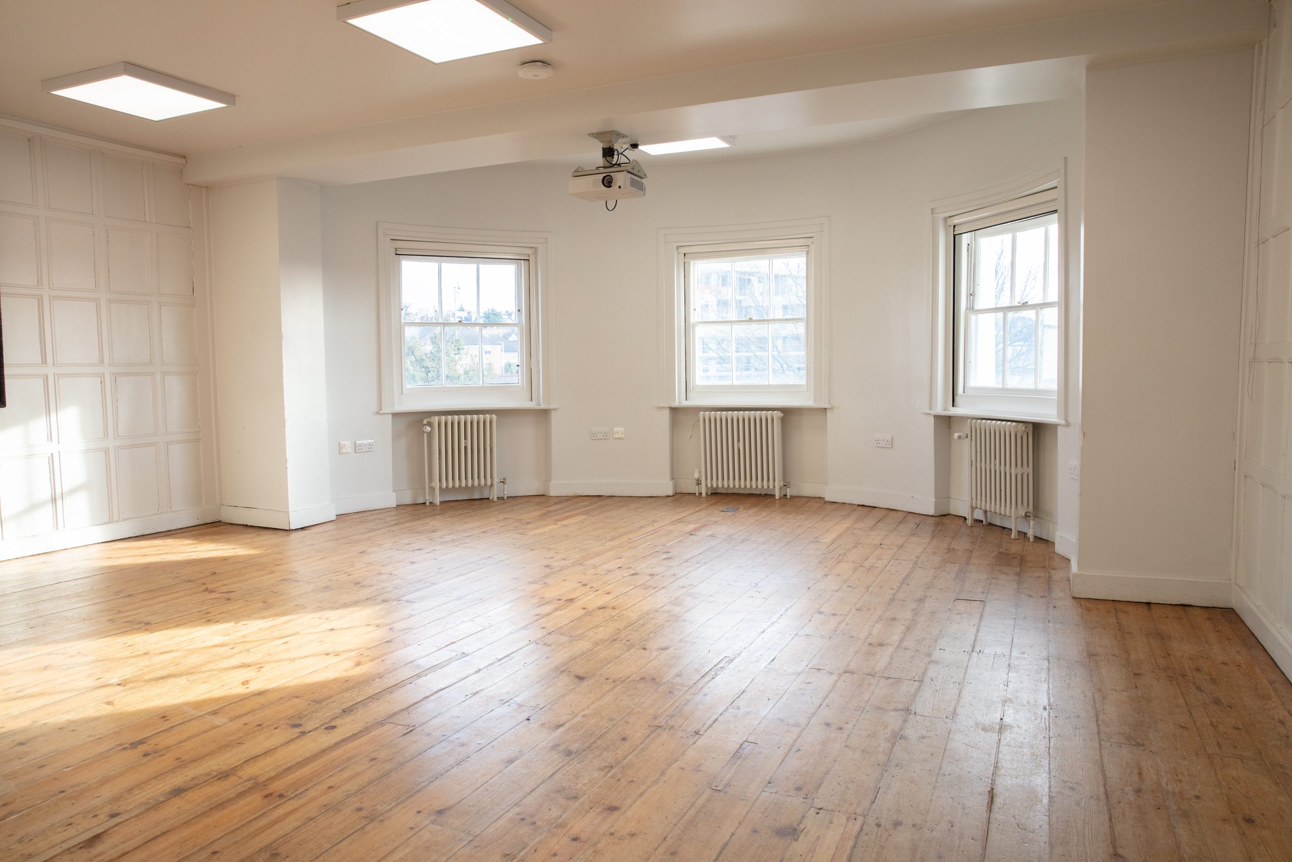 The Acanthus room at William Morris Gallery. The floors are wooden and we have three windows to the right, with sun shining through them. The walls of the room are painted white.