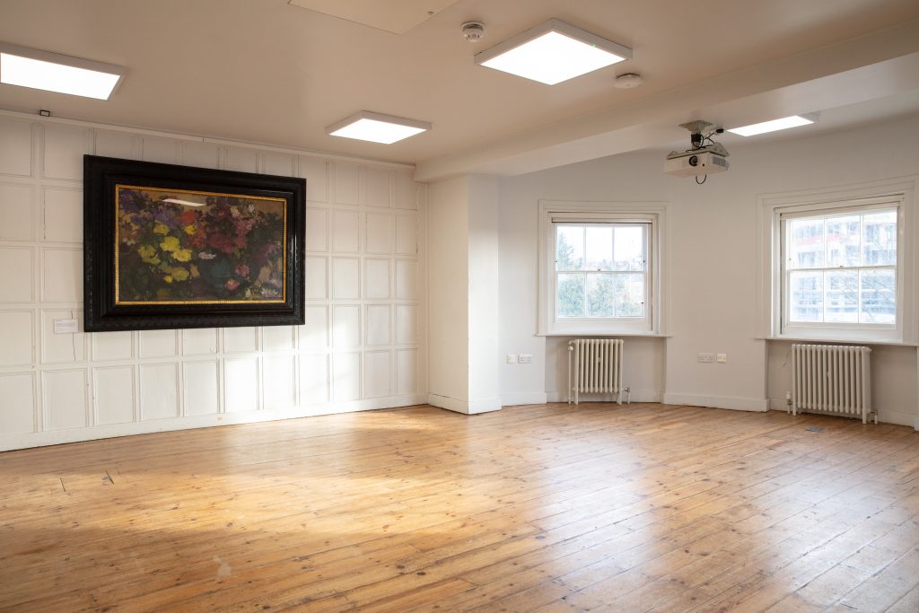 The Acanthus Room at William Morris Gallery. We see an empty room with sun shining in through the windows. The floors are wooden with a painting displayed.