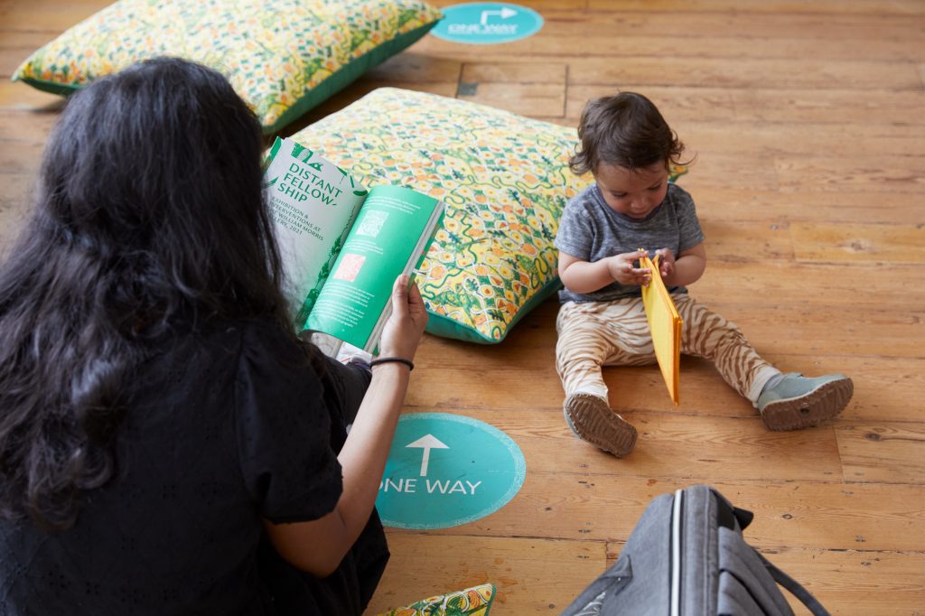 A small child sits on the floor reading, accompanied by its mother. There are cushions on the floor around them.