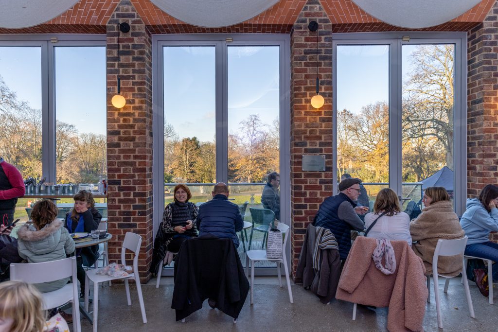 Image of visitors sitting in the Cafe at William Morris Gallery. The William Morris Garden and Lloyd Park can be seen through the windows.