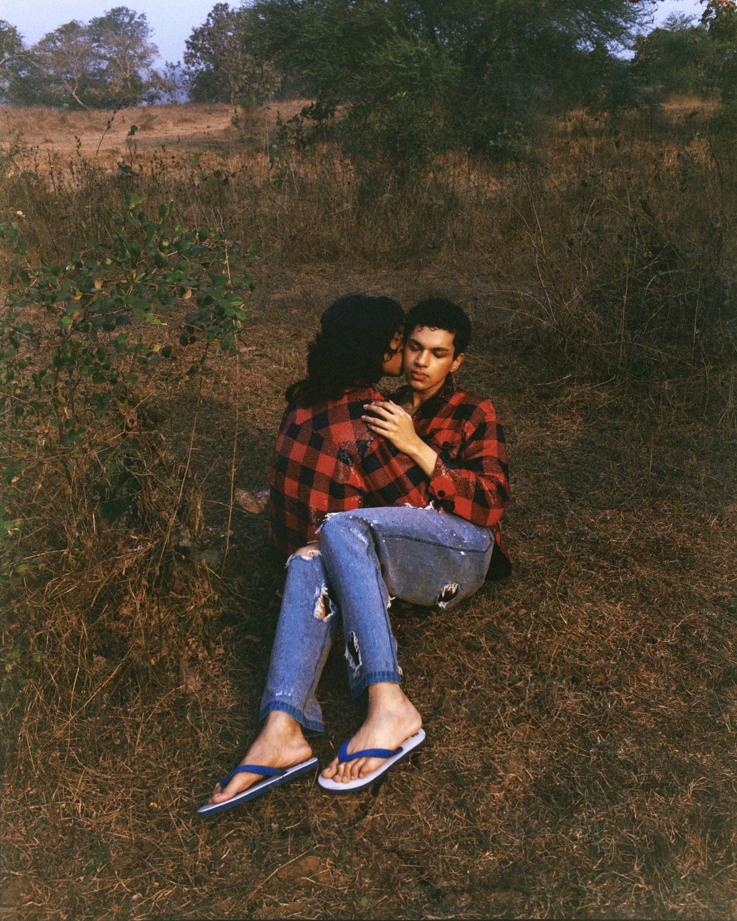 Image of two men embracing. They are sitting on the grass and the face of one person is visible. They are both wearing red and black checkered shirts and the figure to the right is wearing sequined jeans. He also wears blue flip flops. This is an images has a film-like quality.