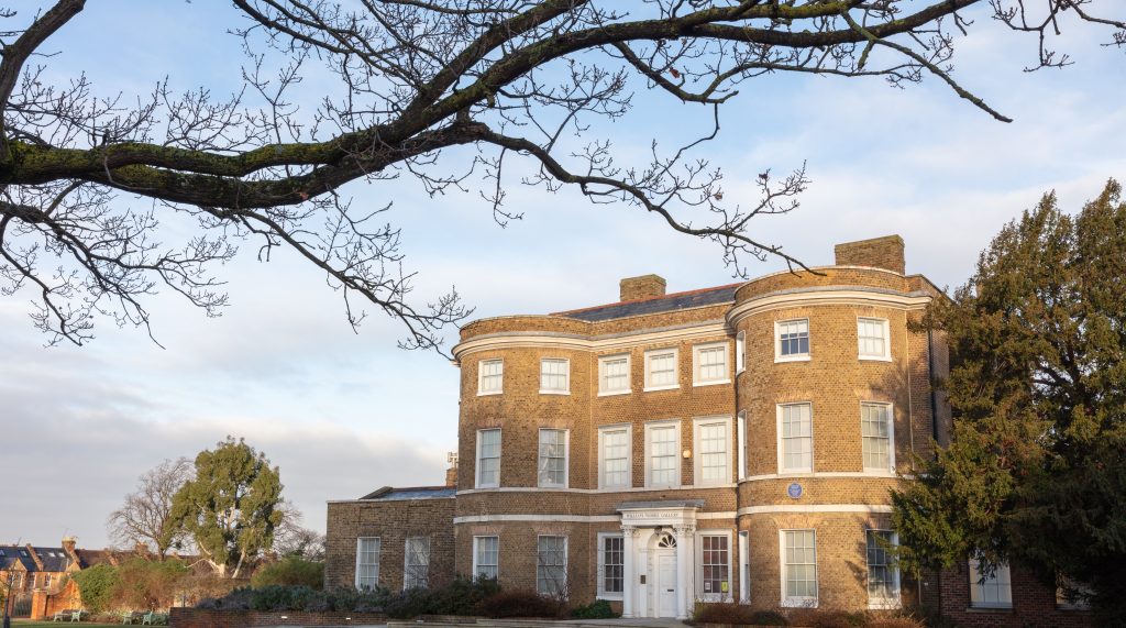 An exterior image of William Morris Gallery. There is a tree branch in the foreground and a tree is also visible to the left of the building.