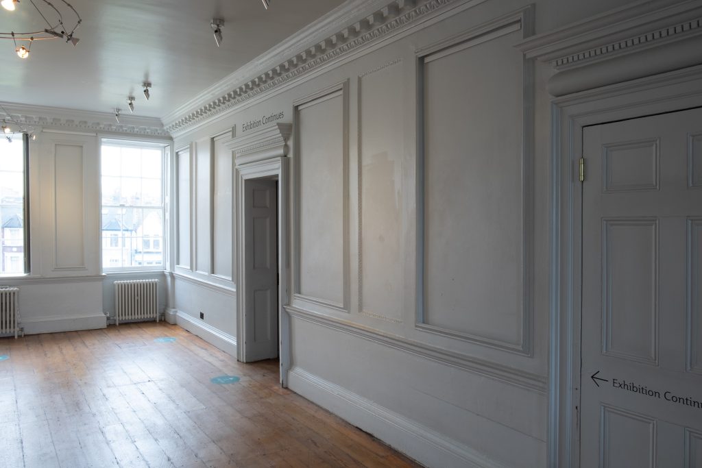 Image of the upstairs landing at William Morris Gallery, with doors leading to the exhibition spaces. Walls are painted white, with wooden floorboards.