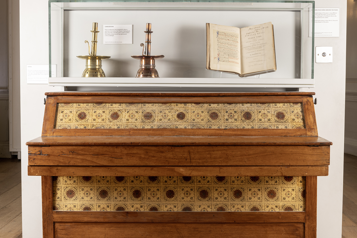 A piano designed by Morris & Co, with candlesticks and an open book displayed above.