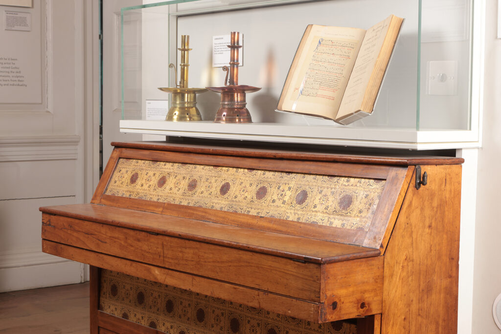 A piano designed by Morris & Co. Books and candlesticks are displayed on top of the piano.
