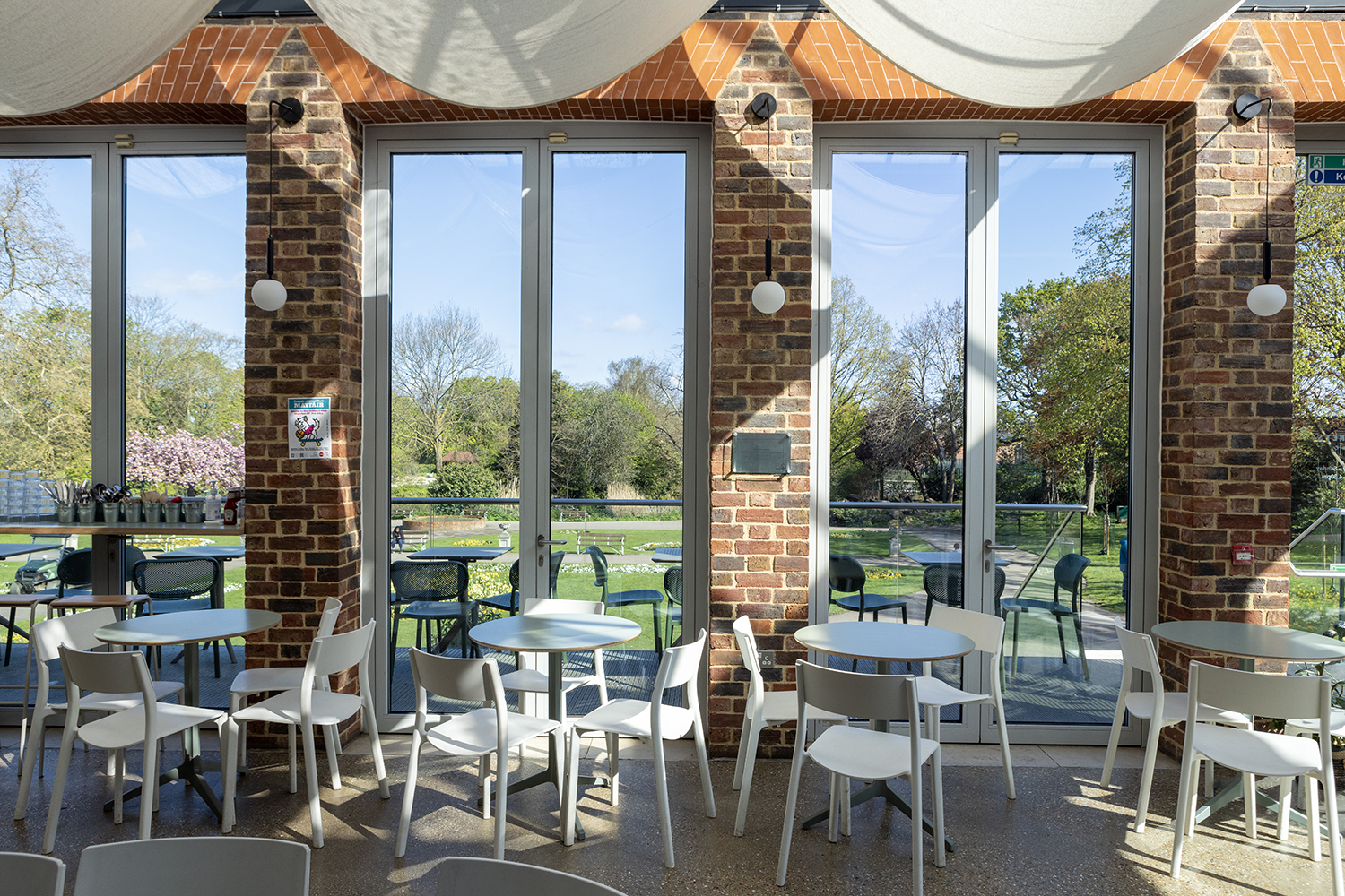 Image of the Cafe at William Morris Gallery on a sunny day. The sun is shining into the space from the ceiling and windows. We see tables, chairs and a counter to the left of the image.
