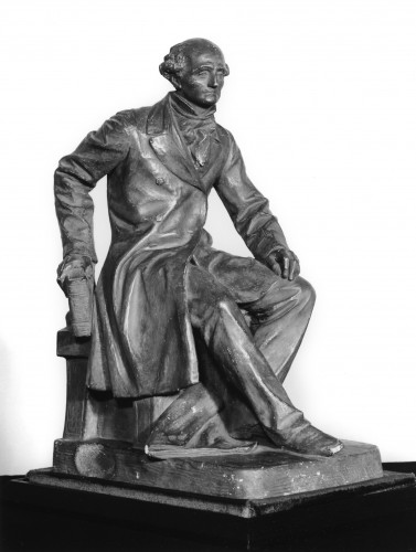 Seated man in cravat, coat and trousers, on low stool with cushion, right hand resting on knee, left hand holding a  resting on back of stool at left.