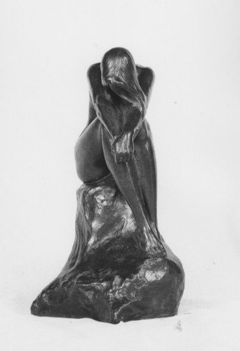 Nude girl seated on a rick her head and shoulders bent forward with her arms resting outstretched on her knees and her hands clasped. Her long hair drapes over her extended arms. Front view.