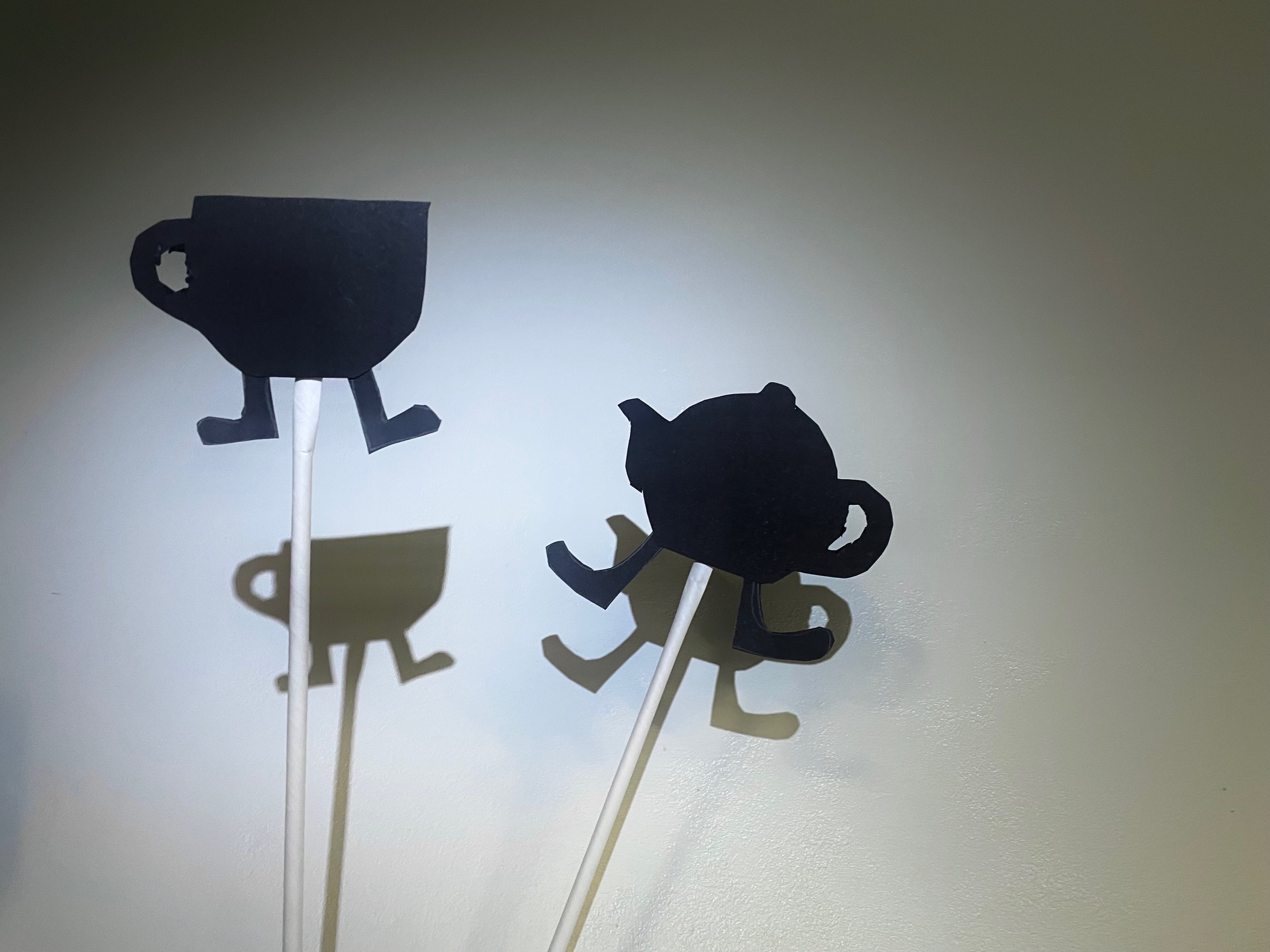 Two shadow puppets in black against a white background. A teapot and a cup, each with feet.