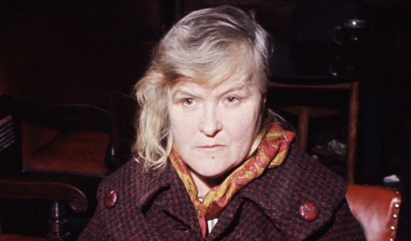 Close up on the face of a woman dressed in an orange scarf and brown coat. She looks severe.